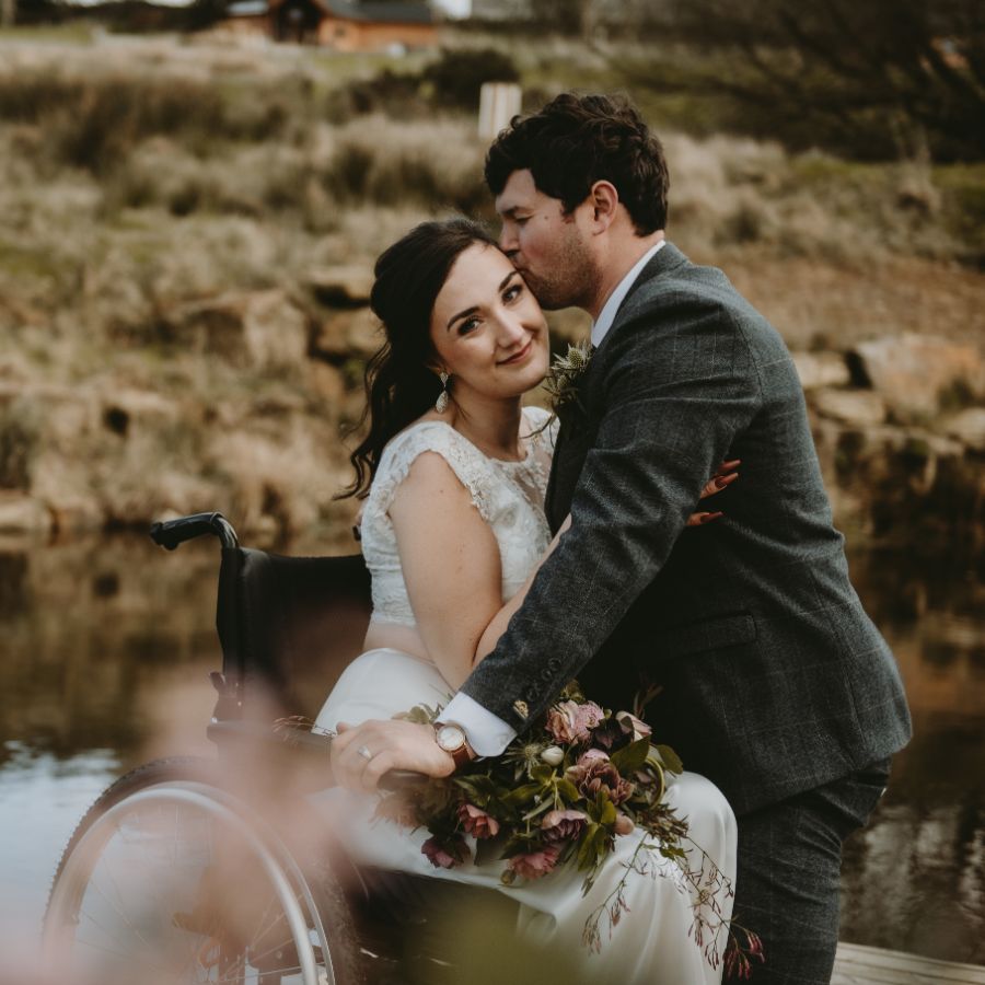 Couple cuddle next to a lake, bride is in a wheelchair